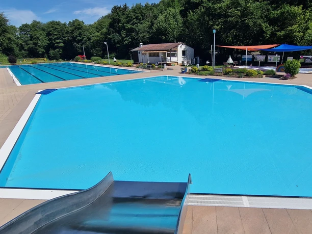 Waldfreibad Valdorf mit blauem Wasser, umgeben von Bäumen, Wasserrutsche im Vordergrund.
