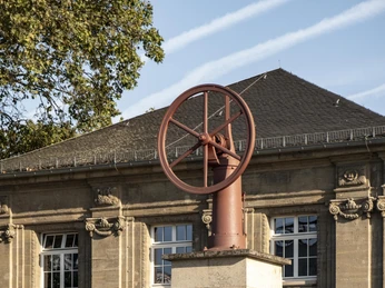Otto- und Langen-Denkmal Das Otto- und Langen-Denkmal zeigt ein Denkmal aus Stein mit einer Darstellung eines Schwungrads auf einem Podest, platziert vor einem historischen Gebäude, das von Bäumen umgeben ist. Fahrräder sind neben dem Denkmal abgestellt.The Otto and Langen memorial is a stone monument depicting a flywheel on a pedestal, placed in front of a historic building surrounded by trees. Bicycles are parked next to the monument.