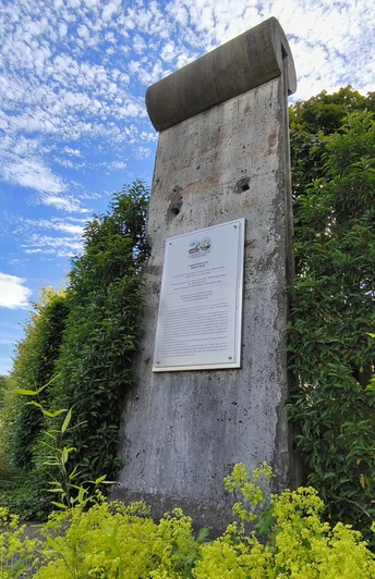 Ein originales Mauerstück der Berliner Mauer, umgeben von grüner Vegetation und einem Infobereich.