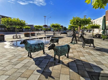 Bronzeskulpturen von Schafen und einem Hirten umgeben einen kleinen Wasserspielplatz am Henkenplatz.