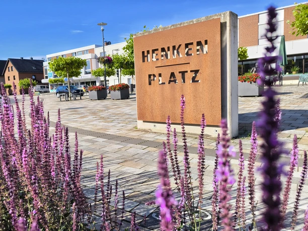 Henkenplatz in Hövelhof mit lila Blumen im Vordergrund und modernen Gebäuden sowie Bäumen im Hintergrund.