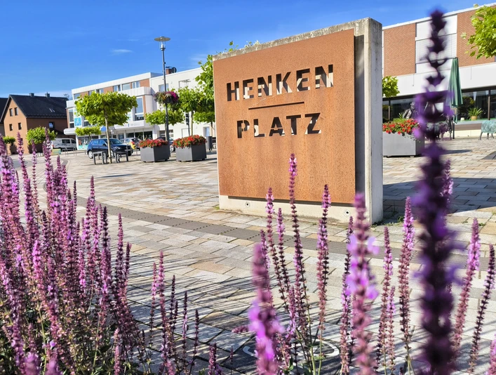 Henkenplatz in Hövelhof mit lila Blumen im Vordergrund und modernen Gebäuden sowie Bäumen im Hintergrund.