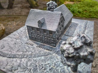 Alter Hövelhof Bronzemodell des Alten Hövelhofs, umgeben von stilisierten Bäumen auf einem Sockel im Freien.