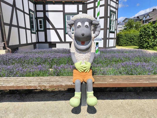 Hoevelhof-Schnucki_Figur_02-CC_BY_SA-Tourist_Information_Hoevelhof.jpg Plüschige Schafskulptur mit orangefarbener Hose sitzt lächelnd vor einem Fachwerkhaus in Hövelhof.