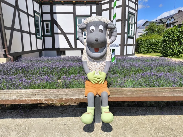 Hoevelhof-Schnucki_Figur_02-CC_BY_SA-Tourist_Information_Hoevelhof.jpg Plüschige Schafskulptur mit orangefarbener Hose sitzt lächelnd vor einem Fachwerkhaus in Hövelhof.