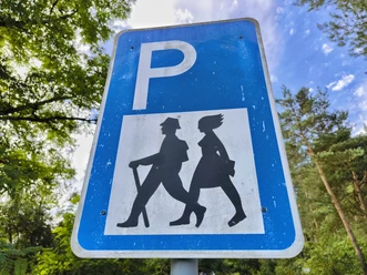 Wanderparkplatz Hövelhof Parkschild mit Wanderermotiv in grüner Umgebung, Himmel im Hintergrund, an Ort Hövelhof positioniert.