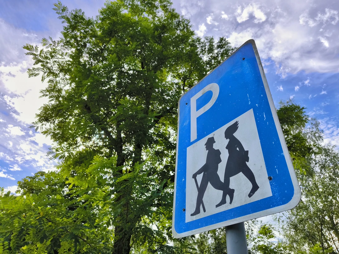 Blaues P-Schild mit Wanderersymbol, umgeben von grünem Laub unter wolkigem Himmel.