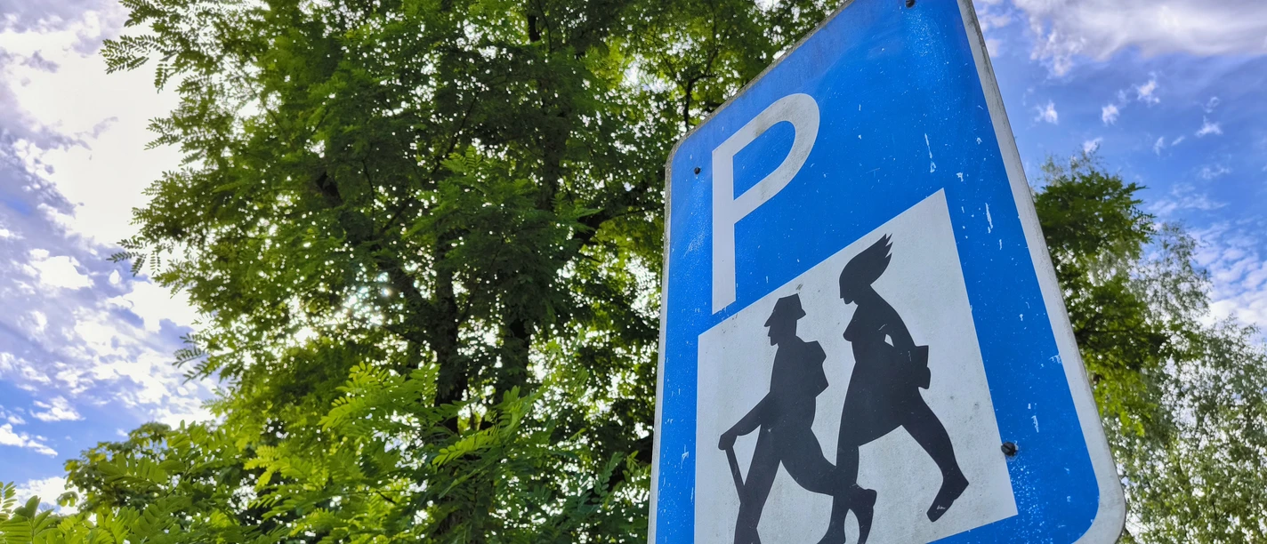 Blaues P-Schild mit Wanderersymbol, umgeben von grünem Laub unter wolkigem Himmel.