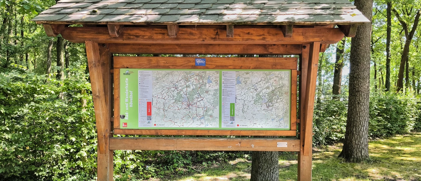 Holztafel vor grünen Bäumen, zeigt Wanderkarten des Gebiets Hövelhof mit Wegen und Routen.
