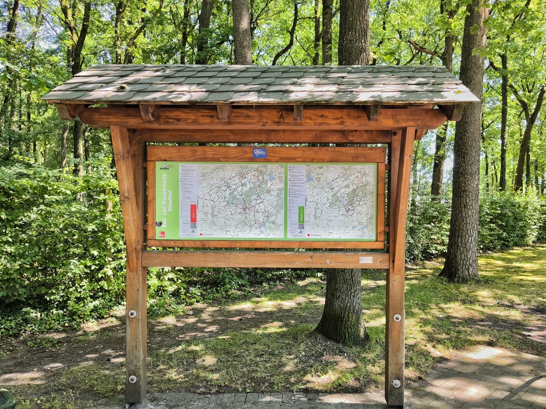 Wanderparkplatz Hövelhof Holztafel vor grünen Bäumen, zeigt Wanderkarten des Gebiets Hövelhof mit Wegen und Routen.
