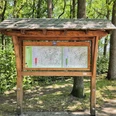 Wanderparkplatz Hövelhof Holztafel vor grünen Bäumen, zeigt Wanderkarten des Gebiets Hövelhof mit Wegen und Routen.