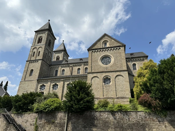 St. Lucia Immekeppel Romanische Kirche mit zwei Türmen und Rosettenfenstern, umgeben von Bäumen, unter blauem Himmel.
