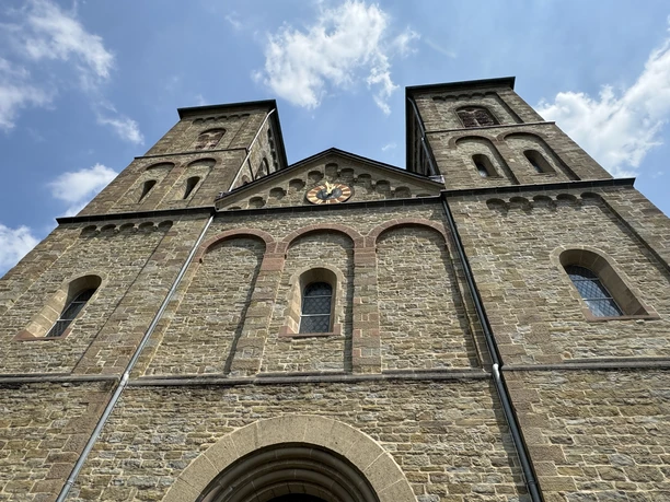 St. Lucia Immekeppel Die Fassade einer historischen Kirche mit zwei markanten Türmen und einem Uhrzifferblatt in der Mitte.