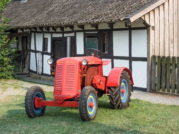 Auf Achse 2018 Roter Oldtimer-Traktor vor traditionellem Fachwerkhaus, auf grünem Gras mit geöffneter Türen.
