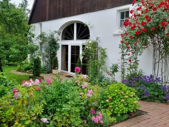Haus.jpg Bild eines charmanten Landhauses mit Blumengarten im Vordergrund, welches von Rosen und Bäumen umgeben ist.