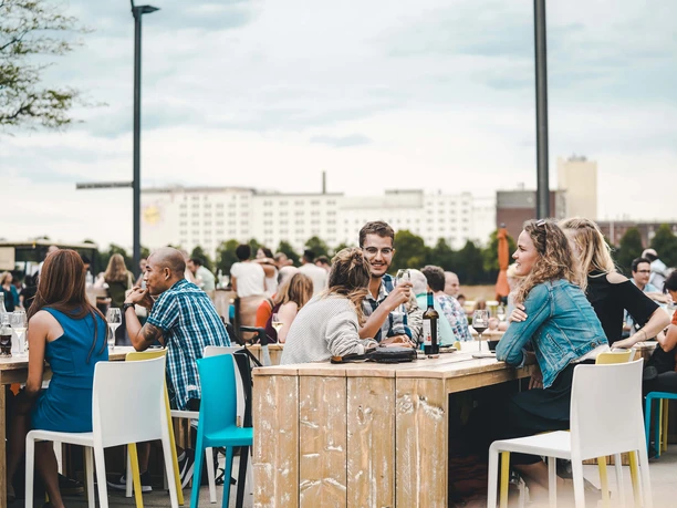 Weinfest am Rhein Menschen sitzen an Holztischen im Freien, genießen Getränke, vor einer urbanen Kulisse.