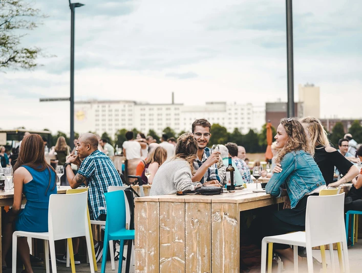 Wine festival on the Rhine Menschen sitzen an Holztischen im Freien, genießen Getränke, vor einer urbanen Kulisse.People sit at wooden tables outdoors, enjoying drinks, against an urban backdrop.
