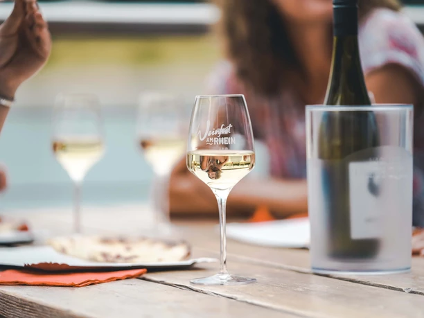 Weinfest am Rhein Das Bild zeigt eine stilvolle Weinszene auf einem Holztisch. Ein Glas Weißwein befindet sich im Fokus, auf dem die Schrift "Weinfest am Rhein" erkennbar ist. Im Hintergrund sind weitere Gläser und unscharfe Umrisse von Personen zu sehen, die eine entspannte und gesellige Atmosphäre vermitteln. Der Tisch ist mit Tellern und einer eingekühlten Flasche bedeckt, was die gesellige Stimmung dieses Weinfestes unterstreicht.