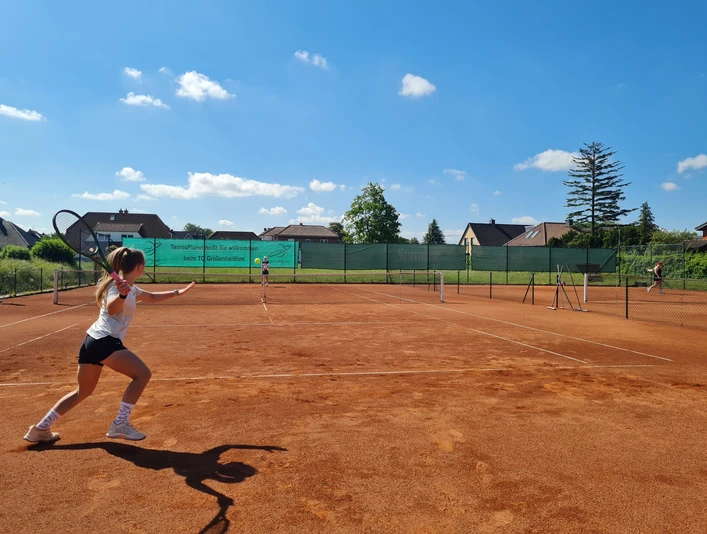 Tennis Club Großenheidorn Eine Frau spielt Tennis auf einem Sandplatz unter blauem Himmel, im Hintergrund sind Häuser.