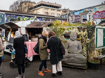 Fair Trödeln Markt in Köln Auf einem urbanen Platz stehen Menschen um einen Imbisswagen. Im Hintergrund sind bunte Graffiti zu sehen, während große Buddha-Statuen zur Ruhe einladen.