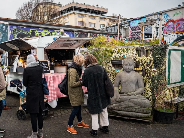 Fair Trödeln Markt in Köln Auf einem urbanen Platz stehen Menschen um einen Imbisswagen. Im Hintergrund sind bunte Graffiti zu sehen, während große Buddha-Statuen zur Ruhe einladen.