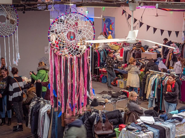 Ein belebter Secondhand-Markt in einer Halle. Menschen stöbern an Ständen mit bunter Kleidung, darunter hängen große, dekorative Traumfänger von der Decke.