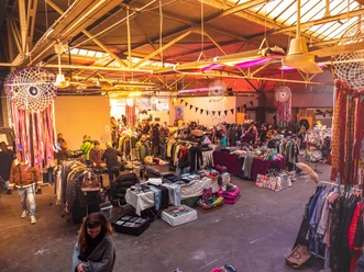 Fair Trödeln Markt in Köln Ein Trödelmarkt in großer Halle: Besucher schlendern zwischen Ständen mit Kleidung, Dekoration und Kunsthandwerk in einer warmen, beleuchteten Atmosphäre.
