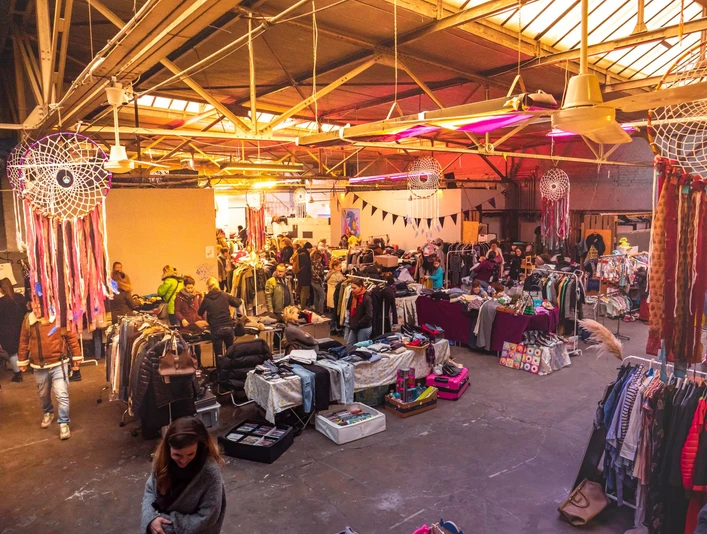 Fair Trödeln Markt in Köln Ein Trödelmarkt in großer Halle: Besucher schlendern zwischen Ständen mit Kleidung, Dekoration und Kunsthandwerk in einer warmen, beleuchteten Atmosphäre.