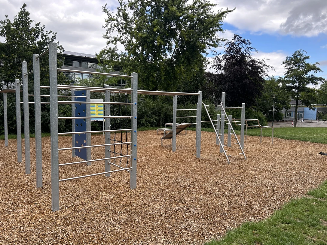 Outdoor-Sportplatz mit Klettergerüsten auf Rindenmulch, umgeben von Bäumen und Rasenfläche.