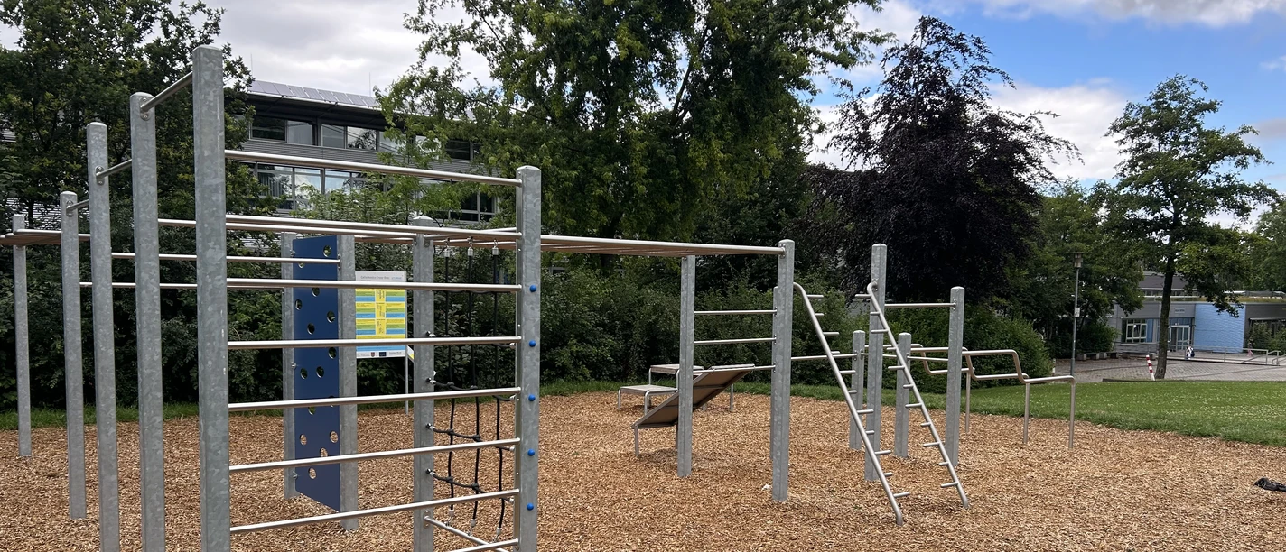 Büren_Sportparcours.jpeg Outdoor-Sportplatz mit Klettergerüsten auf Rindenmulch, umgeben von Bäumen und Rasenfläche.