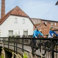 Radfahren im Osnabrücker Land Zwei Fahrradfahrer überqueren eine Brücke vor historischen Backsteingebäuden.