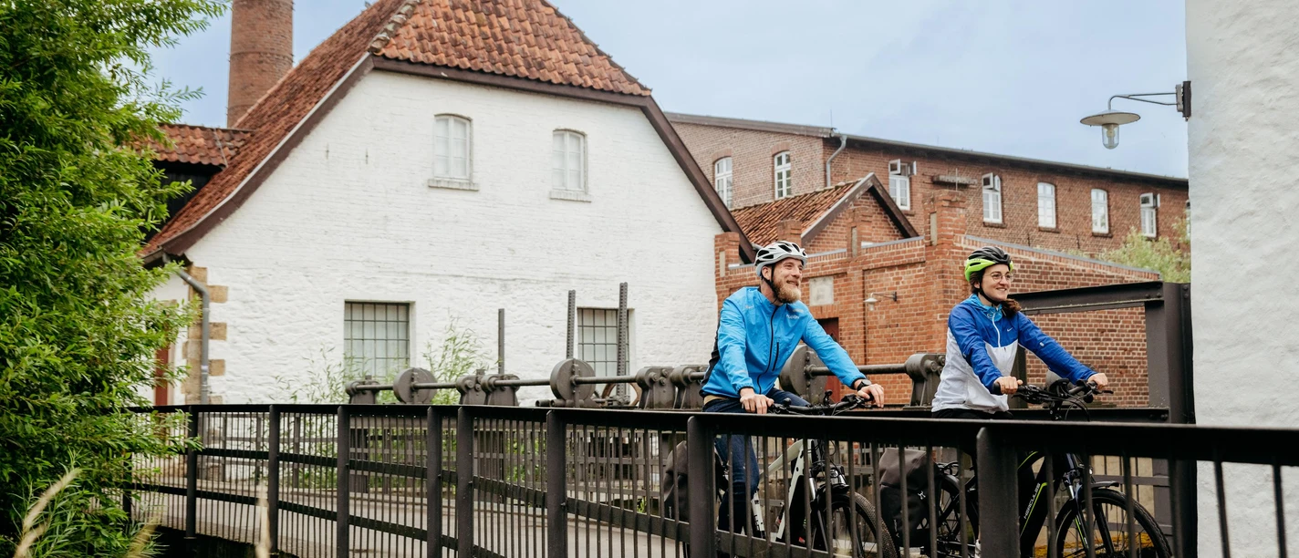 Radfahren im Osnabrücker Land Zwei Fahrradfahrer überqueren eine Brücke vor historischen Backsteingebäuden.