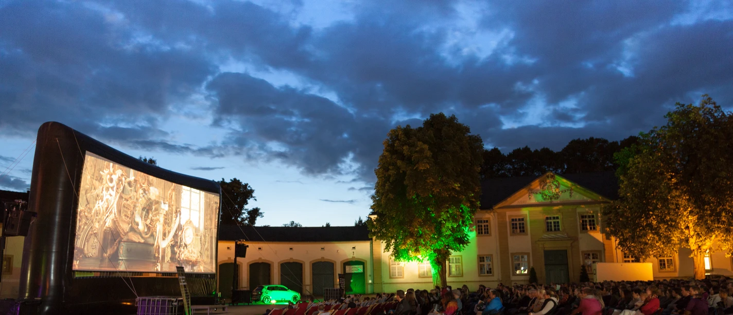 Open-Air-Kino bei Nacht mit Leinwand und zuschauenden Menschen vor alter Gebäudekulisse.