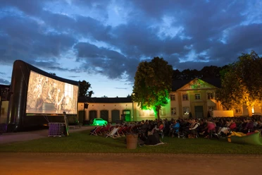 Sommernachtskino.jpg Open-Air-Kino bei Nacht mit Leinwand und zuschauenden Menschen vor alter Gebäudekulisse.