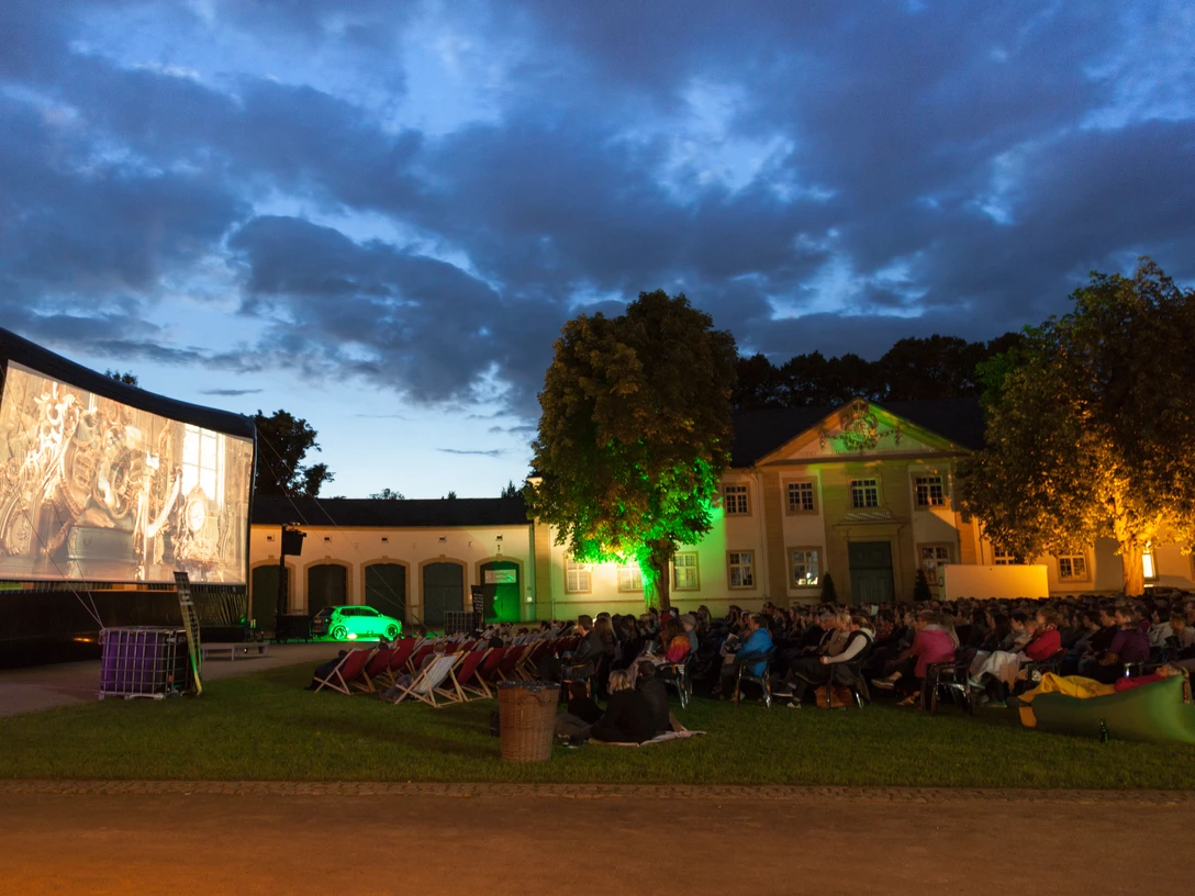 Sommernachtskino.jpg Open-Air-Kino bei Nacht mit Leinwand und zuschauenden Menschen vor alter Gebäudekulisse.