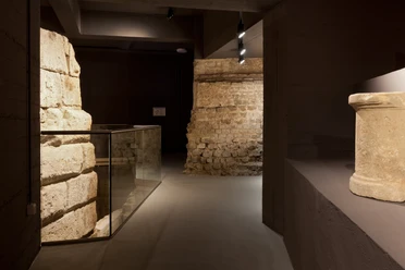Ubiermonument Im Bild sind historische Mauerwerke aus dem antiken Ubiermonument in Köln zu sehen. Im Vordergrund ein Zylinder, dahinter hell beleuchtete Steinwände, die die archäologische Atmosphäre unterstreichen.