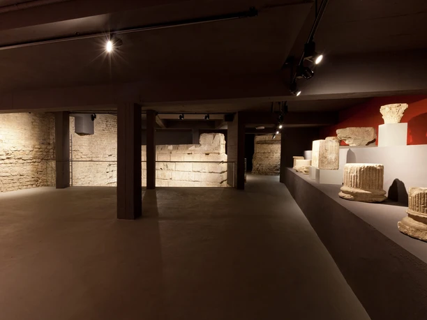 Ubiermonument A museum room with dim lighting shows ancient stones and the remains of columns in a barren cellar.