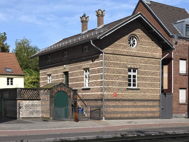 Römergrab Weiden Das Bild zeigt ein altes, gut erhaltenes Backsteinhaus mit schmalen Fenstern und gemauertem Schornstein. Der Eingangsbereich ist durch einen gewölbten Torbogen gekennzeichnet, der in einem ebenfalls aus Backstein gebauten Flügel mündet.