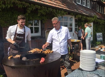 Taurus Weidefleisch ©Emsland Tourismus GmbH Zwei Männer grillen Fleisch und Brotstücke an einer großen Feuerplatte vor einem Bauernhaus.