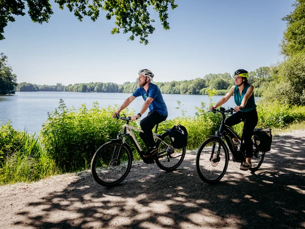 Zwei Radfahrer radeln bei sonnigem Wetter auf einem Pfad entlang eines malerischen Sees.