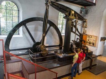 Dauerausstellung im MIK Zwei Personen betrachten eine historische Dampfmaschine mit Schwungrad in einem Industriemuseum.
