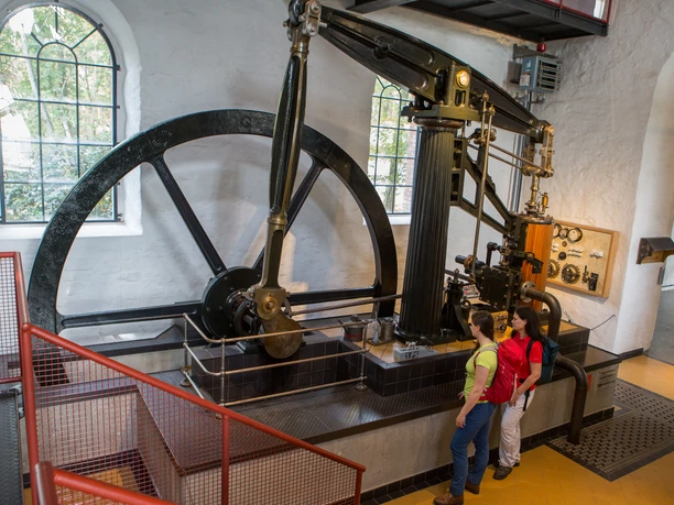 Dauerausstellung im MIK Zwei Personen betrachten eine historische Dampfmaschine mit Schwungrad in einem Industriemuseum.