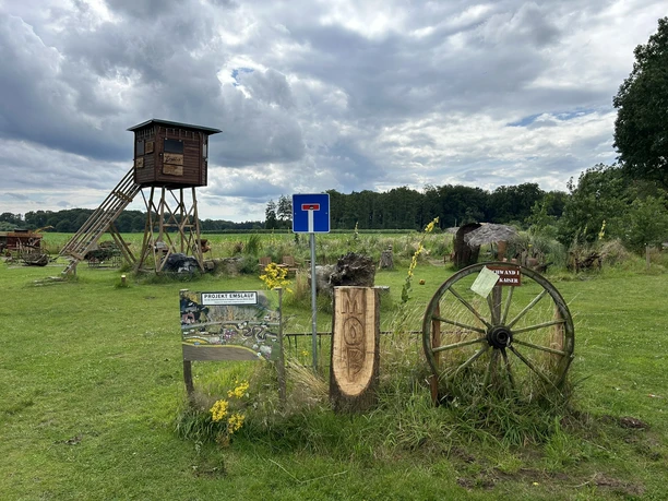 Ems in Kleinformat ©Emsland Tourismus GmbH (2).JPEG Grüner Park mit Aussichtsturm, Holzskulpturen, historischen Wagen und Schautafel unter Wolkenhimmel.