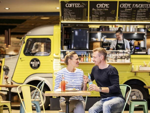 Een jong stel zit gelukkig aan een tafel bij een Franse bistro foodtruck in een markthal.