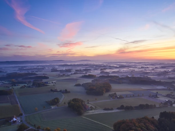Landschaft im Morgennebel mit weiten Feldern und buntem Himmel bei Sonnenaufgang.