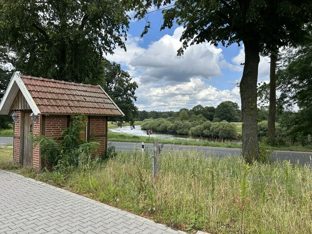 Hofladen Pott-Stopp Lingen Wachendorf ©Emsland Tourismus GmbH (13).JPEG Kleines Backsteinhäuschen am Wegesrand mit Blick auf Fluss, Wiesen und bewölkten Sommerhimmel.