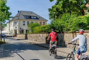 Zwei Radfahrer fahren eine gepflasterte Straße entlang, vorbei an einem gelben zweistöckigen Gebäude.