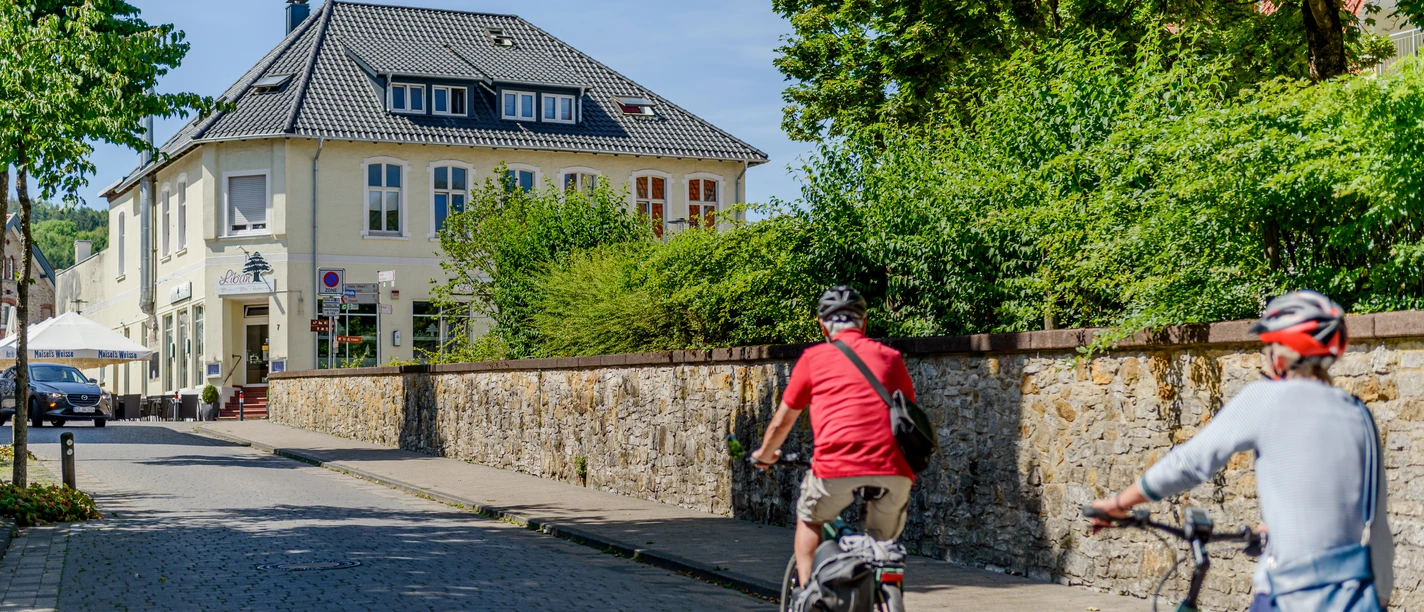 Zwei Radfahrer fahren eine gepflasterte Straße entlang, vorbei an einem gelben zweistöckigen Gebäude.