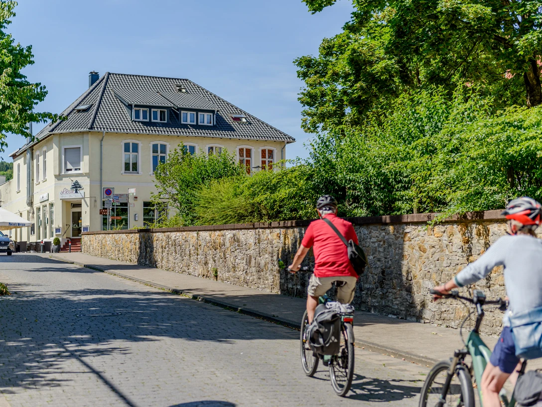 _BBB7394.jpeg Zwei Radfahrer fahren eine gepflasterte Straße entlang, vorbei an einem gelben zweistöckigen Gebäude.