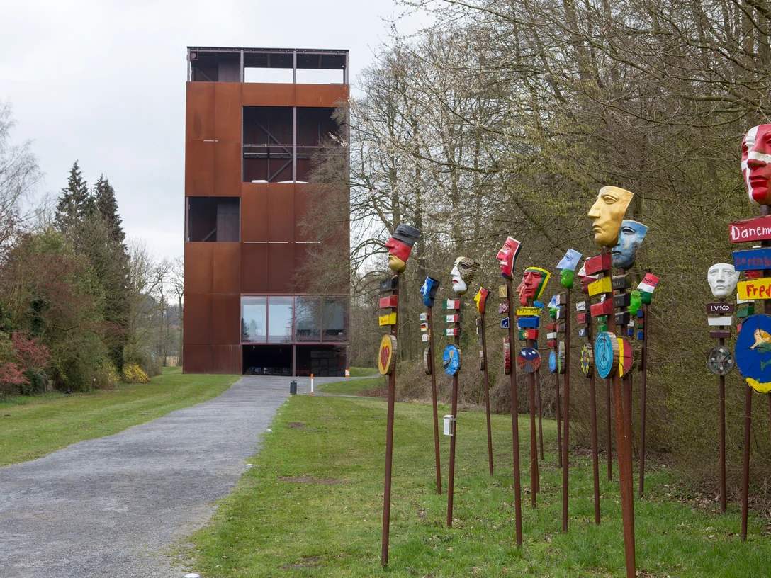 Varusschlacht im Osnabrücker Land - Museum und Park Kalkriese Rostiger Turm neben bunten, bemalten Maskenpfählen auf einem Weg durch Bäume.Rusty tower next to colorful, painted mask poles on a path through trees.Rustent tårn ved siden af farverige, malede maskestolper på en sti gennem træerne.Roestige toren naast kleurrijke, beschilderde maskerpalen op een pad tussen bomen.
